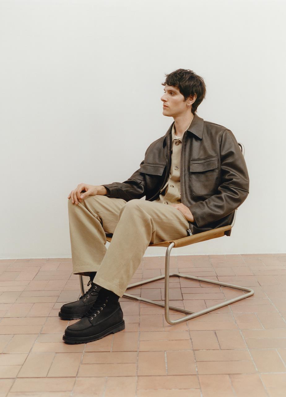 A man in a brown leather jacket sits on a chair, showcasing the off-black Jay Boots in nubuck