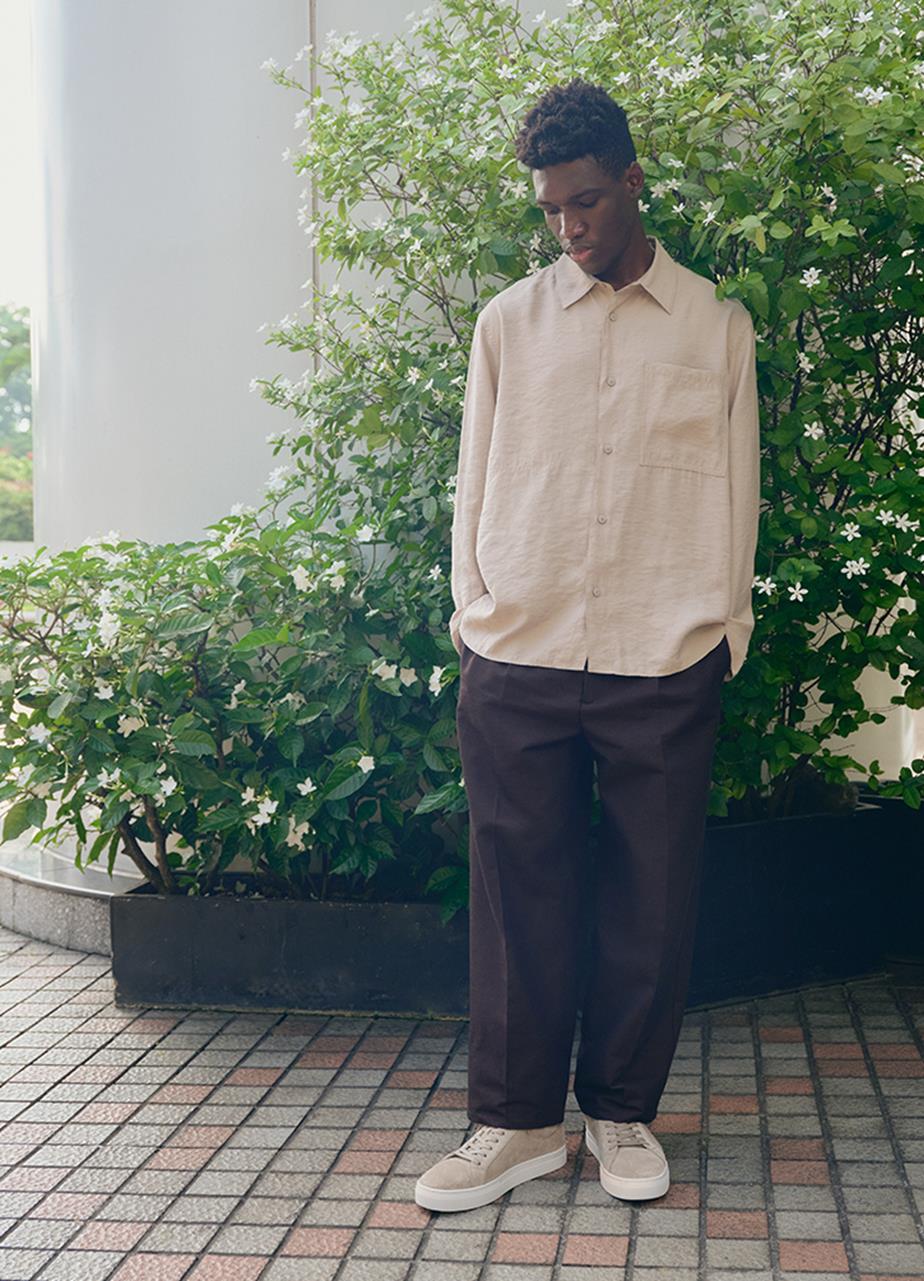 A man stands in front of a large green plant, wearing black pants, sneakers and a casual shirt.