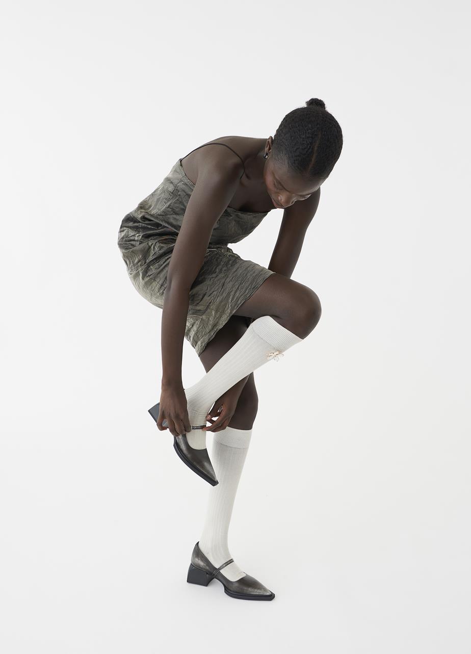 Female model styled in a mid-rise dress and knee-high socks, adjusting the buckle of her silver Mary Jane shoes.