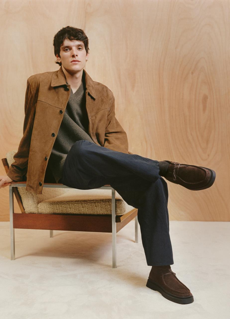 A man in a brown jacket sits on a chair, showcasing the dark brown suede Mike Boots