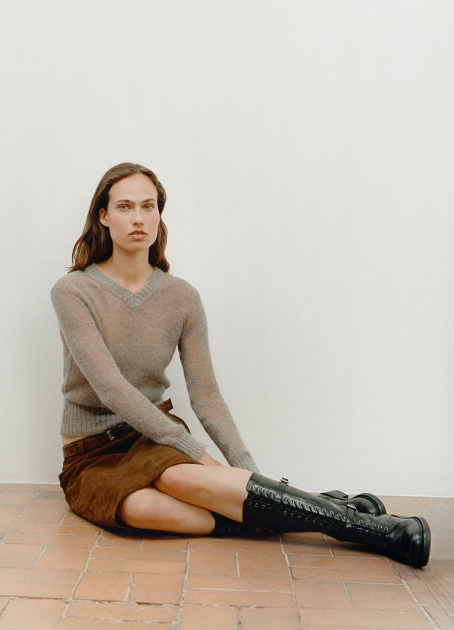 A woman in a long sleeve knit and a brown skirt is sitting on a brick floor, showcasing the Karlie Tall Boots