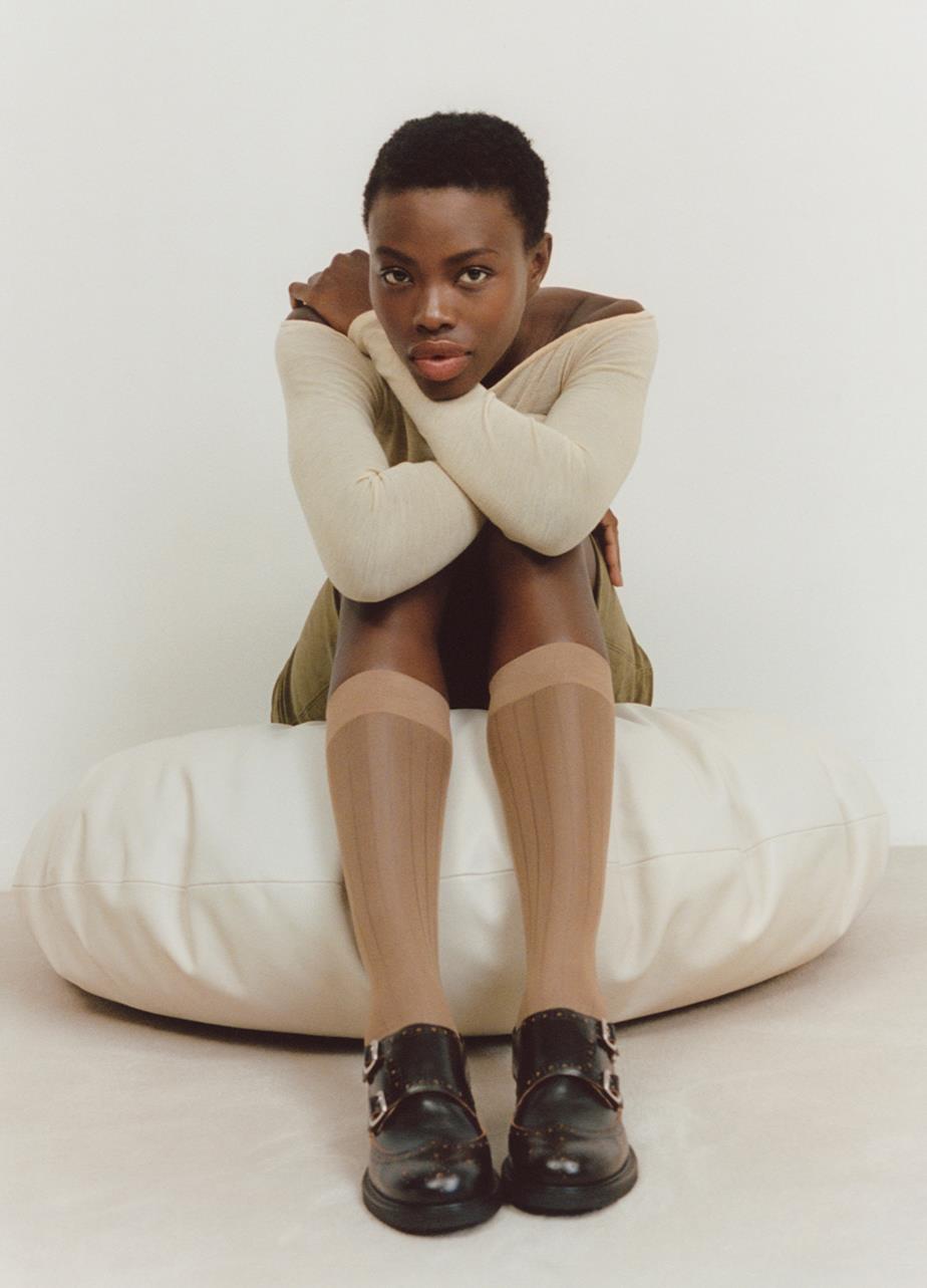 A woman sits on a bean bag, wearing socks and showcasing the black leather Freya Monk Shoes