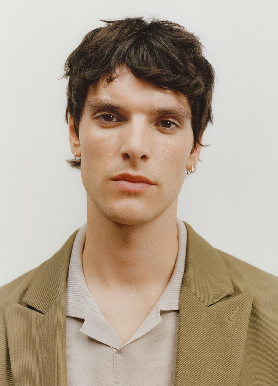 A close-up picture of a mans face with brown hair, a light beige shirt and a green/beige jacket - looking straight in to the camera