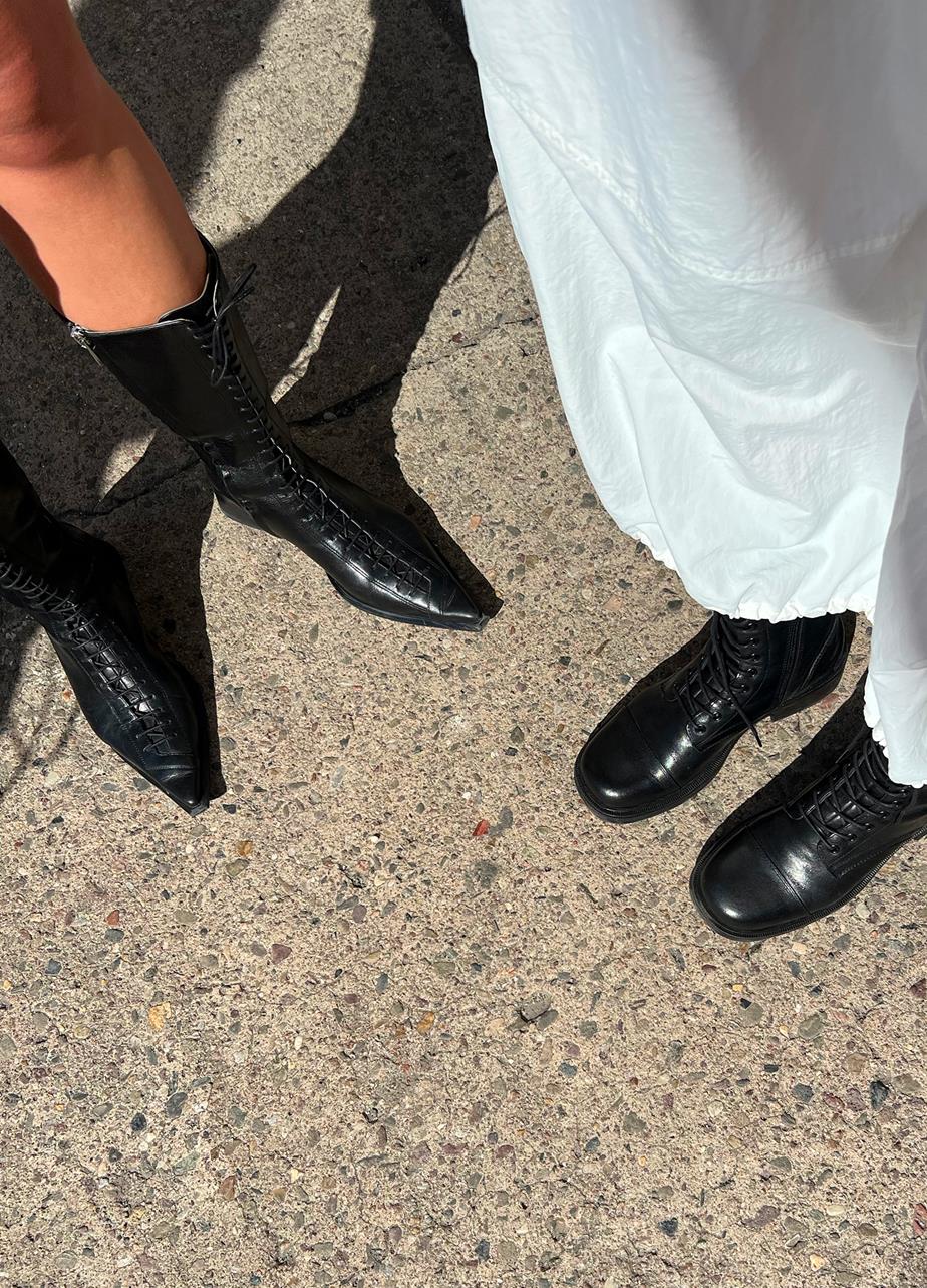 Two individuals in Samria and Karlie black boots standing on a sidewalk, facing each other with a cityscape in the background