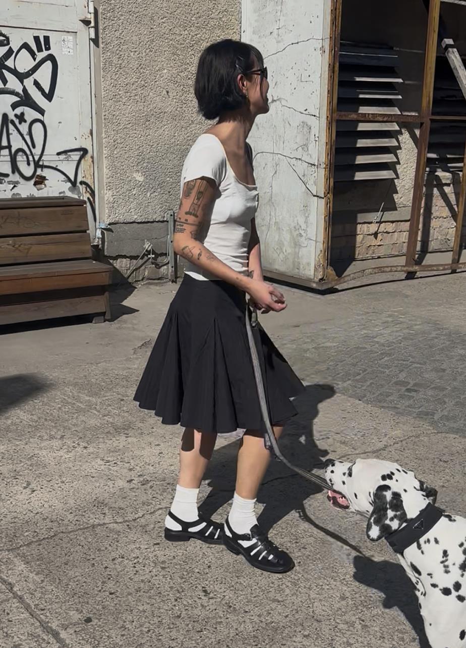 A woman in a black skirt and white shirt walks a Dalmatian dog, wearing black Ellis sandals with white socks.