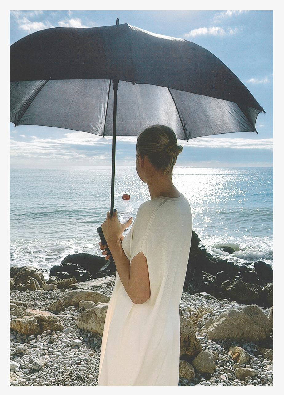 A person is standing and gazing out over the sea, holding an umbrella to shield herself from the sun.