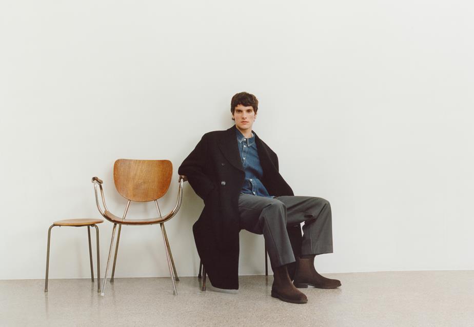 A man with brown hair sitting on a chair in front of a white wall, with two empty chairs next to him, showcasing the Brian Boots he is wearing. 