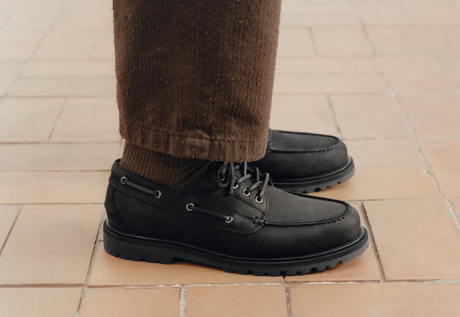 A person with brown pants stands on a brick floor, showcasing the black nubuck Jay Boat Shoe
