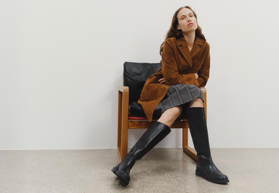 A woman in a brown suede coat sits on a chair, showcasing the Freya Tall Riding Boots in black leather
