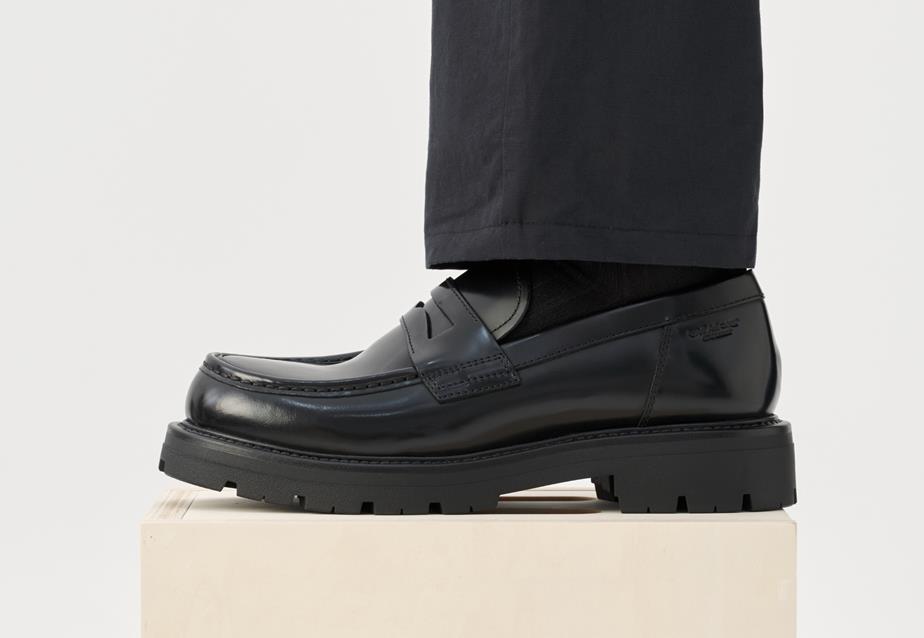 A person with black pants stands on a block, showcasing a profile view of the Cameron Loafer in black