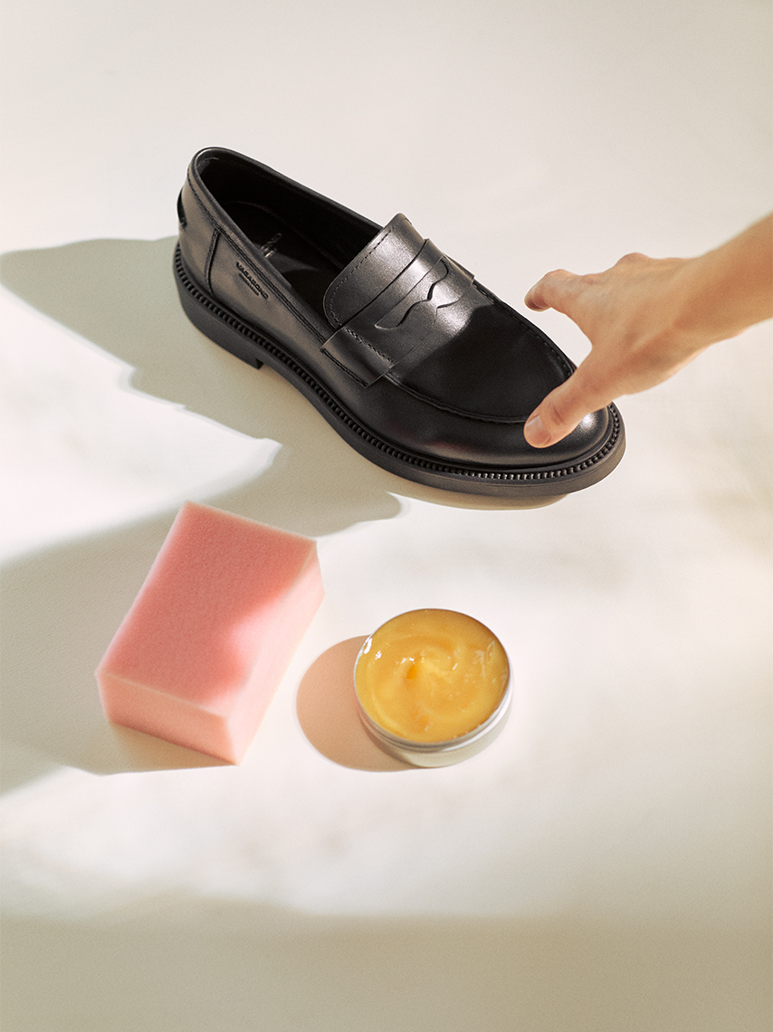 A person picks up a black loafer for polishing, next to shoe wax and a polishing sponge.